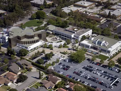 Aerial view of the Cerritos Civic Center showing the Cerritos Library on the left, Cerritos City Hall in the center with a circular plaza, and the Cerritos Sheriff’s Station on the right. Parking lots, landscaped areas, nearby homes, and surrounding commercial buildings are visible.